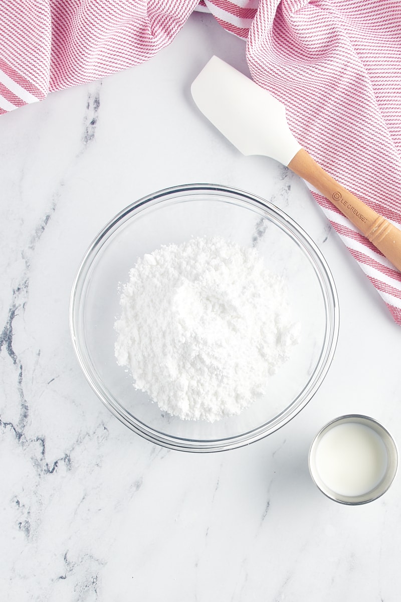 Powdered sugar in a glass bowl with a cup of milk next to it.