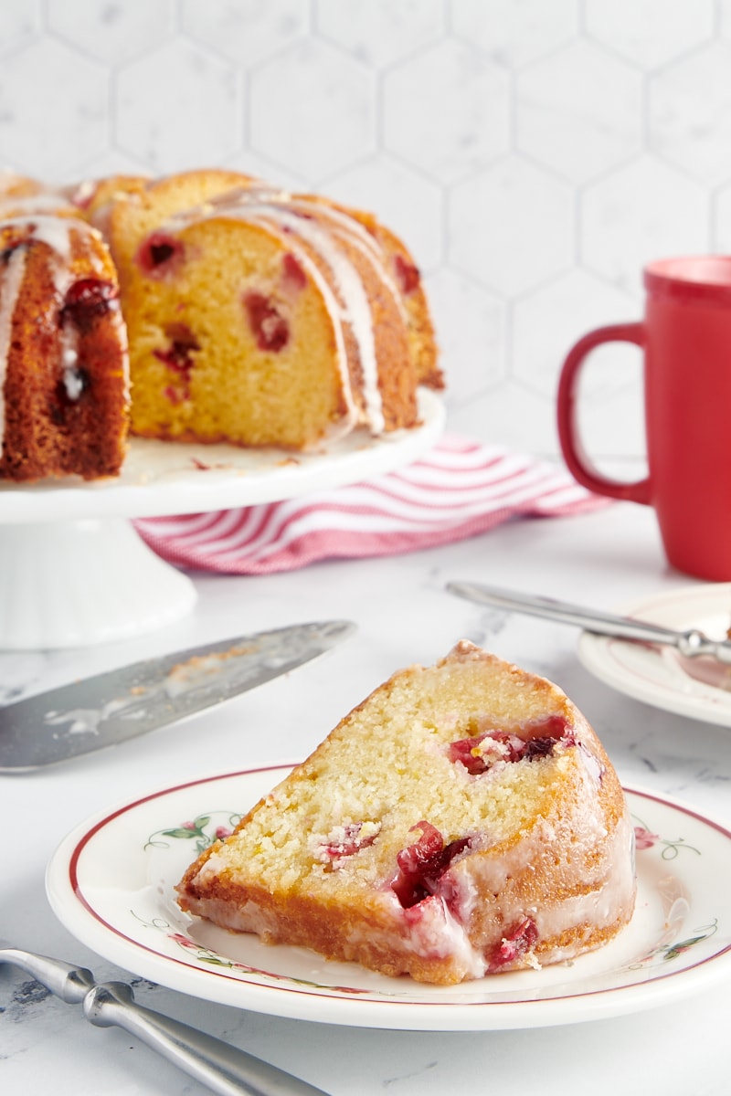 A slice of cranberry bundt cake on a plate with the rest of the cake in the background.