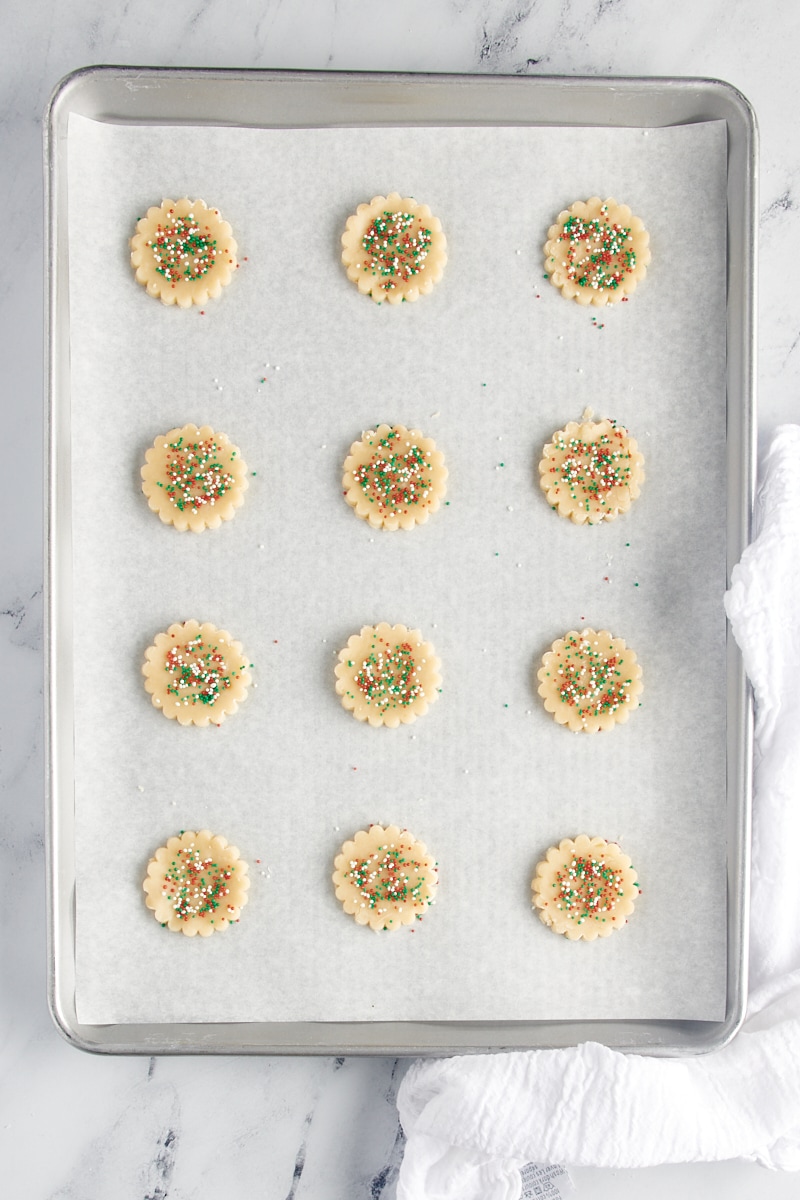 Cutout shortbread cookie dough topped with sprinkles and placed on a parchment-lined baking sheet.