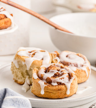 3 cinnamon buns on a plate with a bowl of icing in the background