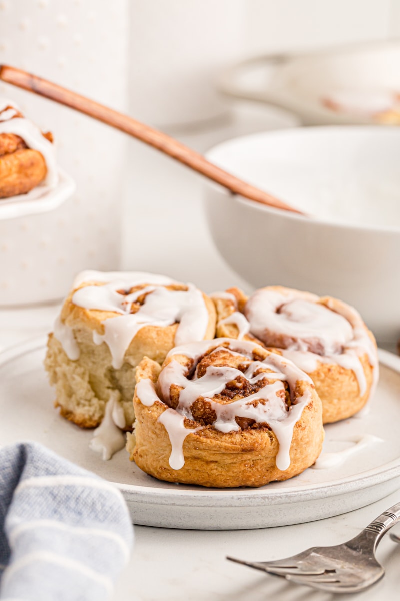 3 cinnamon buns on a plate with a bowl of icing in the background