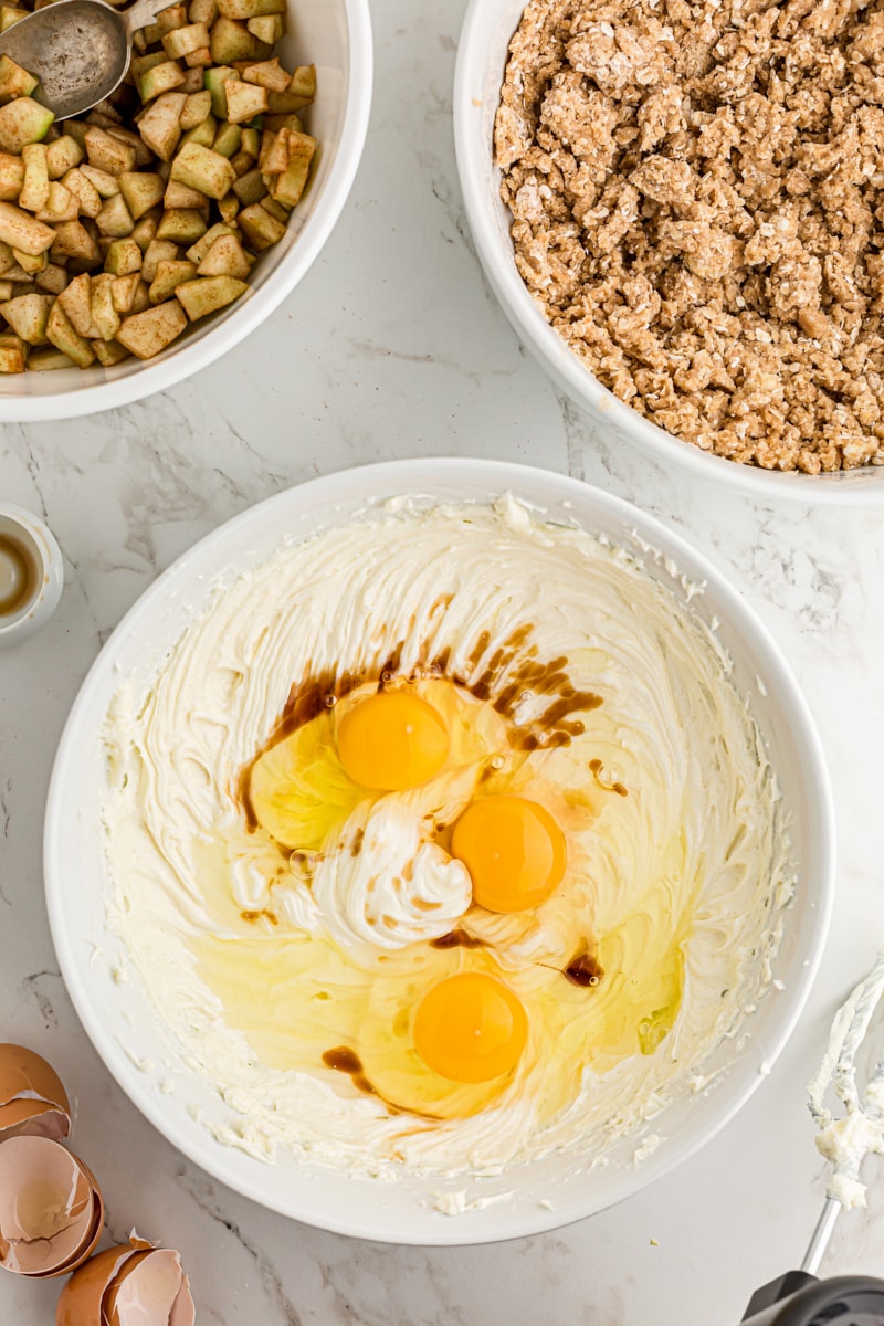 overhead view of eggs and vanilla added to cream cheese and sugar mixture in a white mixing bowl