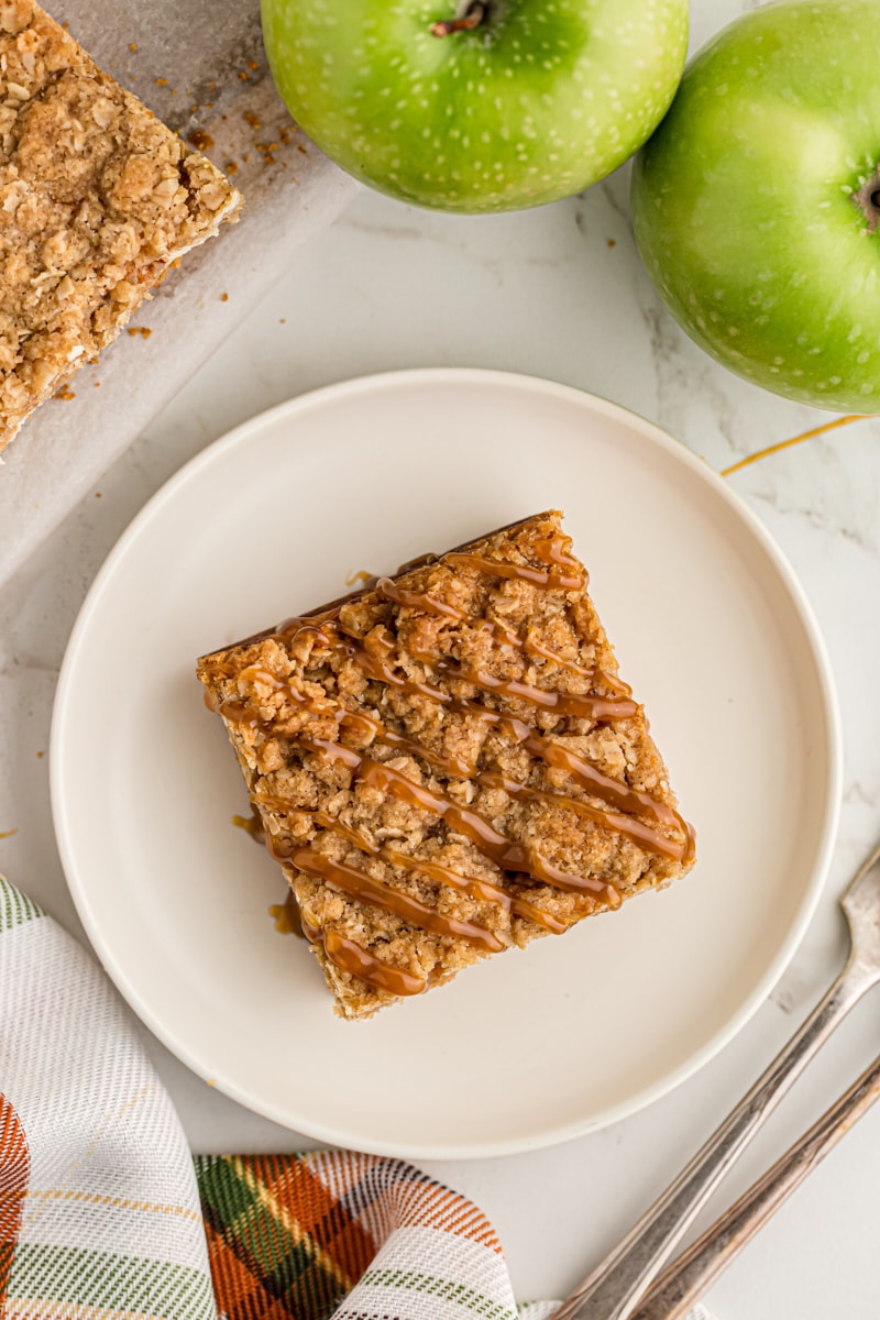 overhead view of a Caramel Apple Cheesecake Bar on a white plate with apples and more bars beside