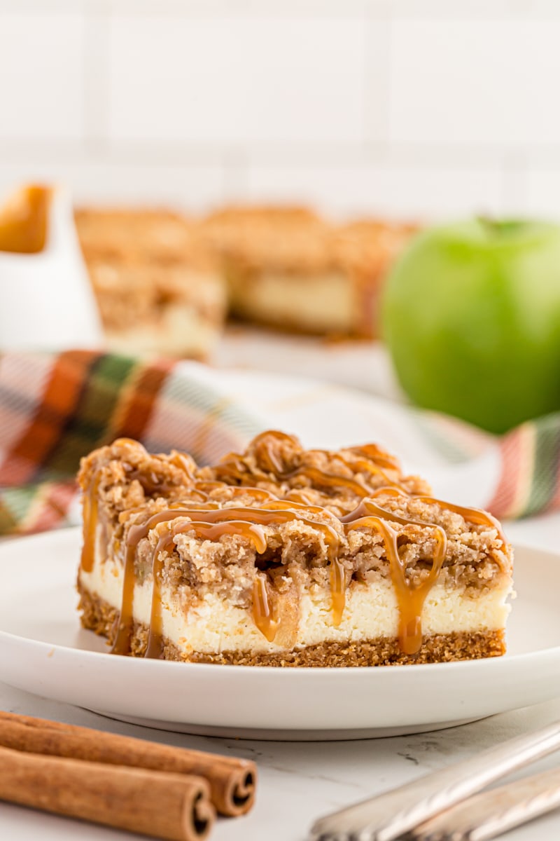 a Caramel Apple Cheesecake Bar on a white plate with more bars on a plate in the background