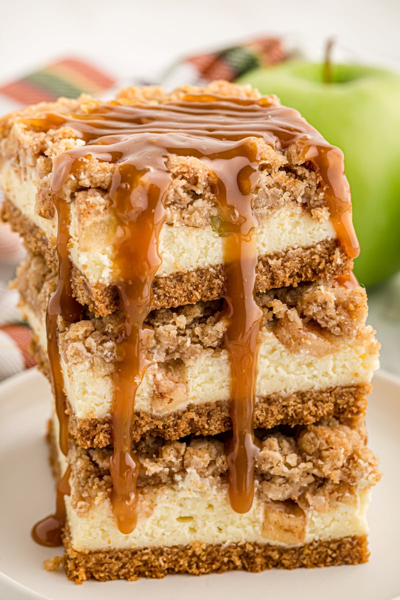 three Caramel Apple Cheesecake Bars stacked on a white plate with caramel sauce drizzled over the top and down the side