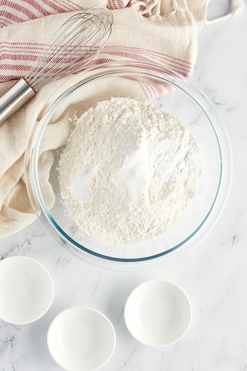 Overhead view of flour, baking powder, baking soda, and salt in a glass mixing bowl.