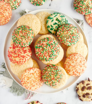 overhead view of sugar cookies piled on a large white plate with more cookies surrounding the plate