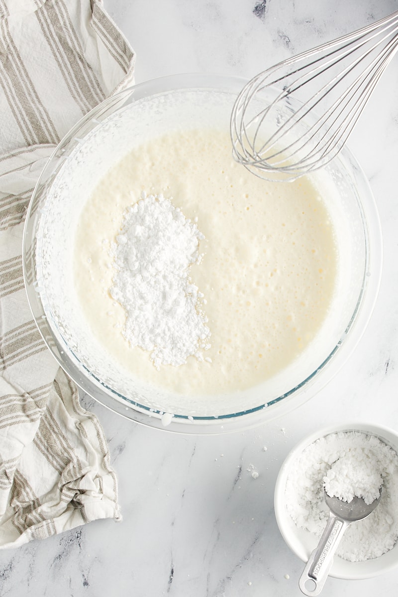 overhead view of confectioners' sugar added to partially whipped cream in a glass mixing bowl