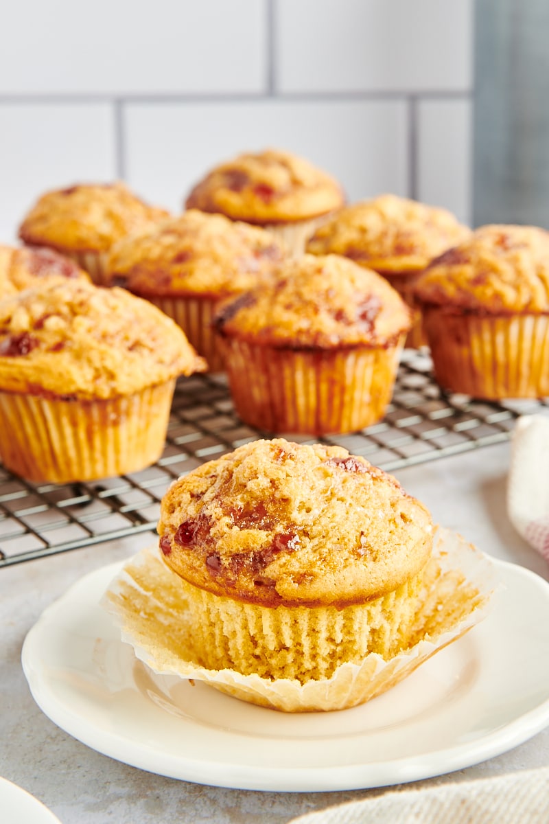 a partially unwrapped Peanut Butter and Jelly Muffin on a white plate with more muffins on a wire rack in the background