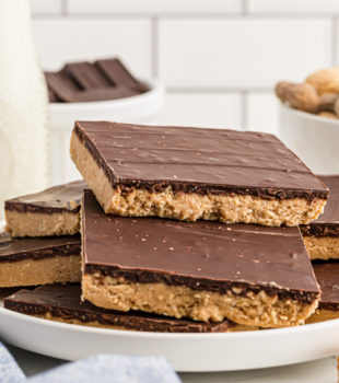 Assorted no-bake peanut butter bars stacked on plate with a bowl of raw peanuts and a jug of milk in the background.