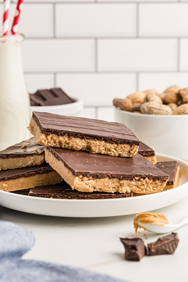 Assorted no-bake peanut butter bars stacked on plate with a bowl of raw peanuts and a jug of milk in the background.