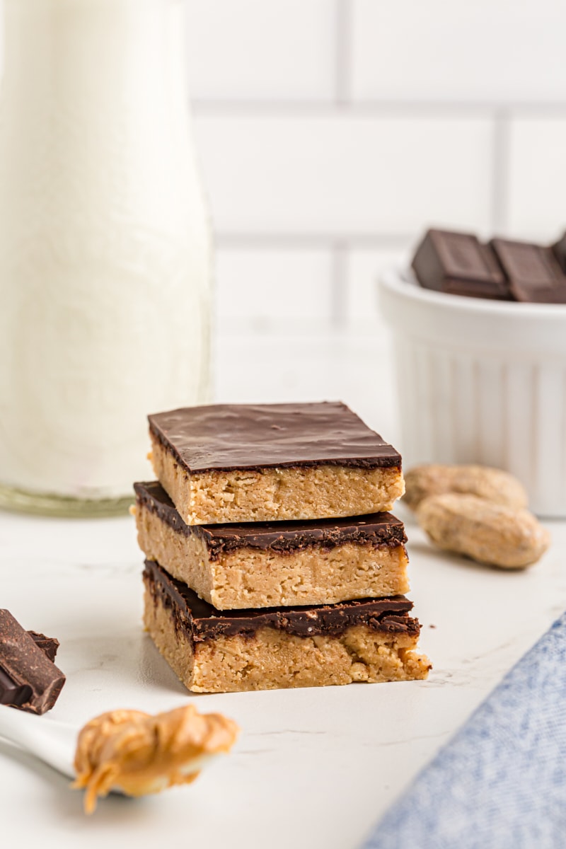 Three no-bake peanut butter bars stacked on a countertop with a bowl of chopped chocolate and a jug of milk in the background.