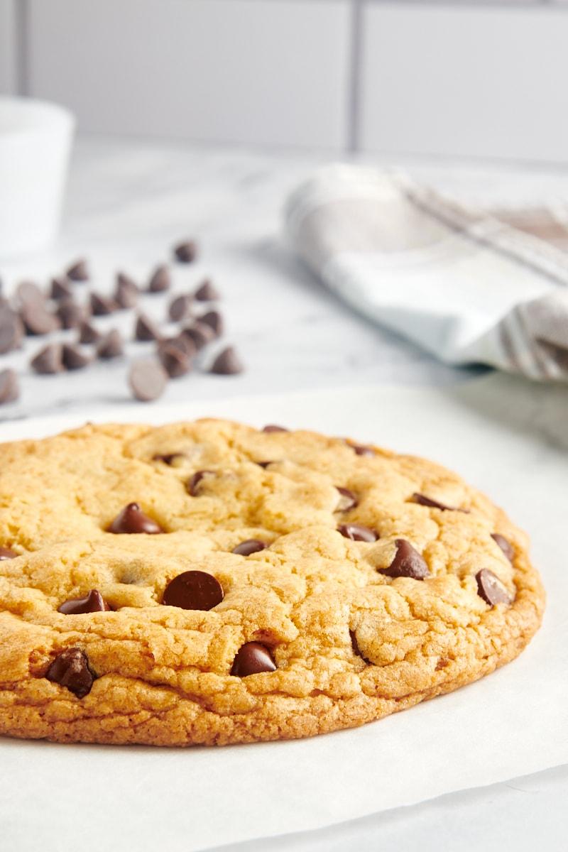 a giant chocolate chip cookie on parchment paper with chocolate chips scattered behind it