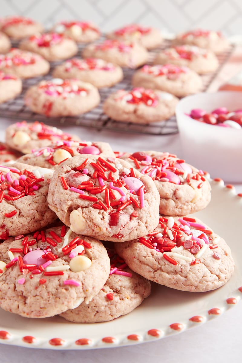 pile of Strawberry M&M Cookies on a red-rimmed white plate with more cookies on a cooling rack in the background