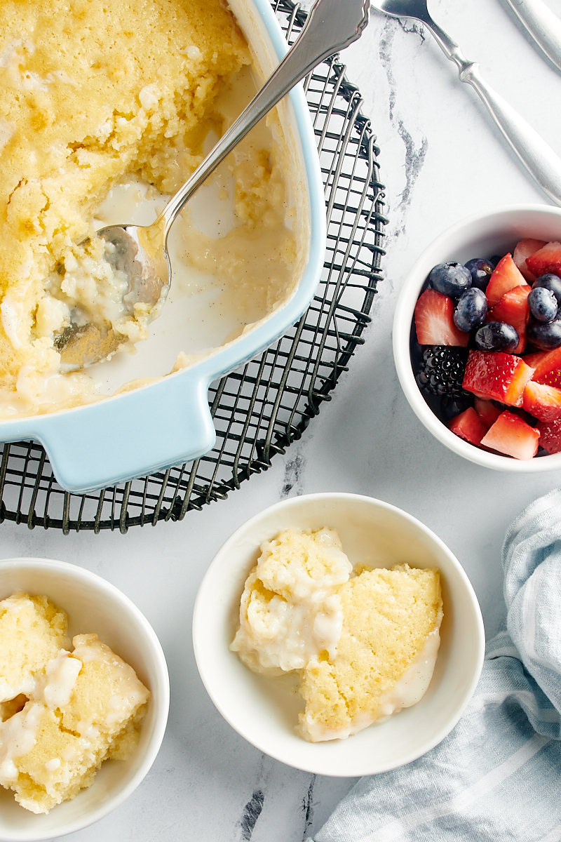 overhead view of two servings of Vanilla Pudding Cake, the remaining cake in a baking pan, and a bowl of mixed berries