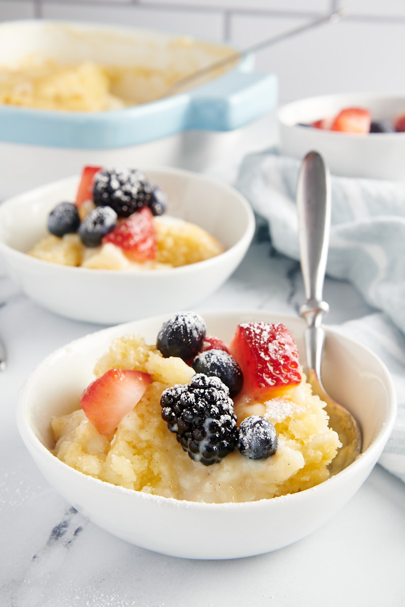 a bowl of Vanilla Pudding Cake served in a white bowl and topped with fresh berries and confectioners' sugar