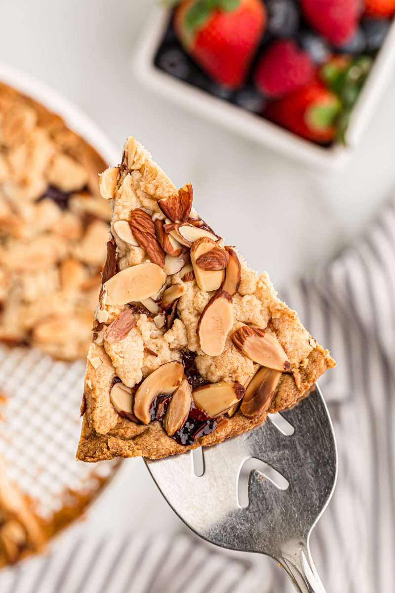 slice of Italian Jam Crostata on a pie server with the remaining tart and a bowl of fresh fruit in the background