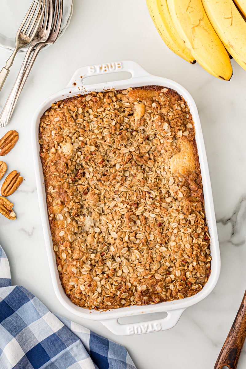 Overhead view of banana bread cobbler in white baking dish, surrounded by bananas, pecans, and forks