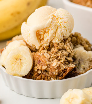 Banana bread cobbler in white dish topped with scoop of vanilla ice cream and banana slices