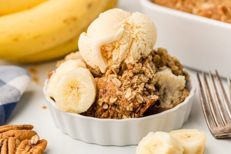 Banana bread cobbler in white dish topped with scoop of vanilla ice cream and banana slices