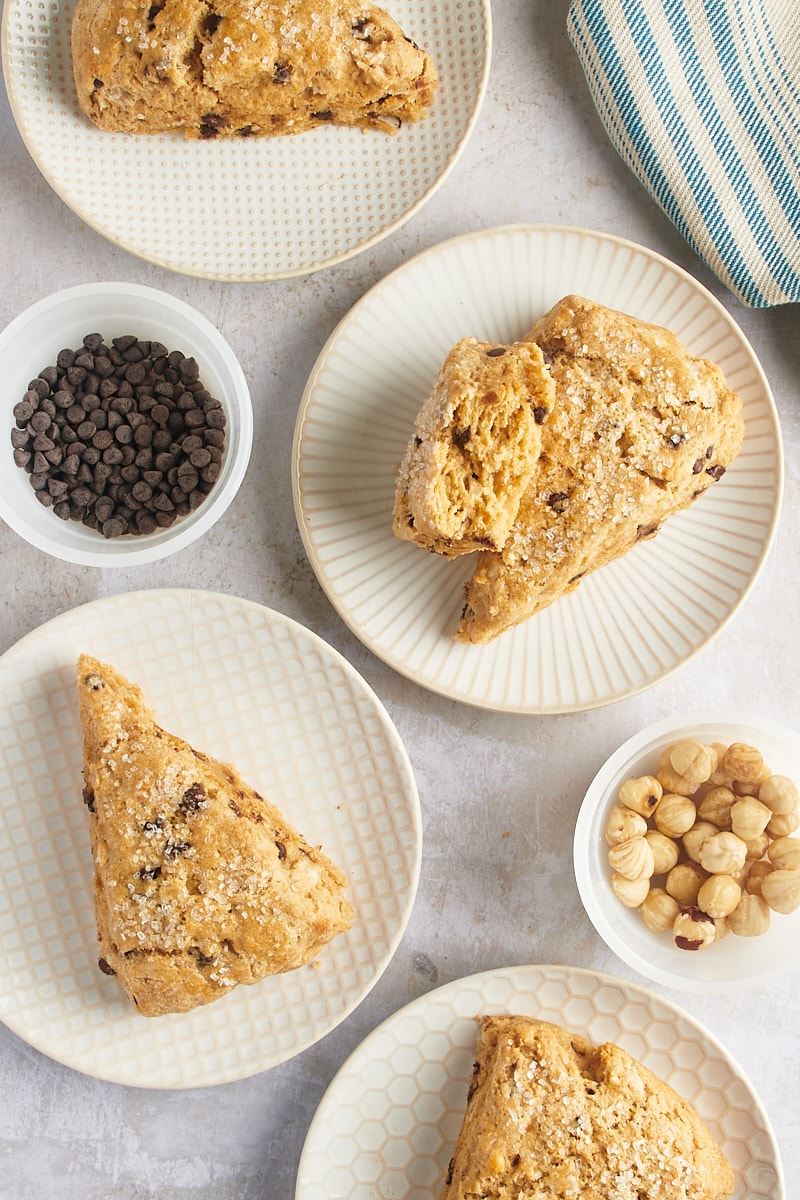 Chocolate chip-hazelnut scones on white patterned plates with bowls of chocolate chips and hazelnuts