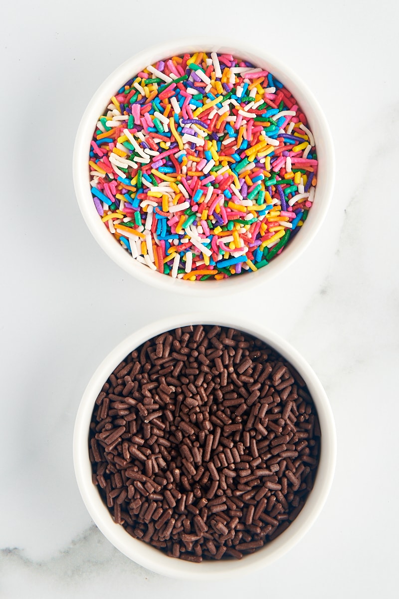 overhead view of rainbow jimmies and chocolate jimmies in white bowls on a marble surface
