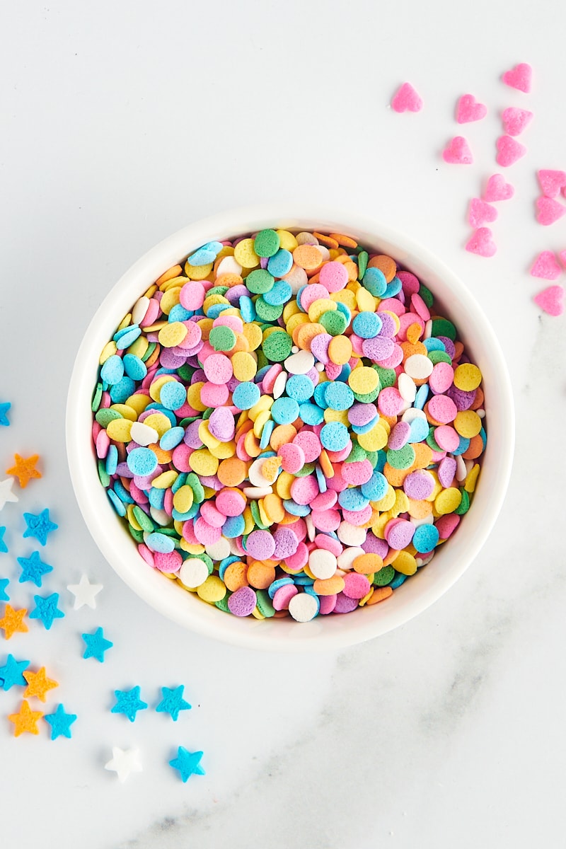 overhead view of quins sprinkles in a white bowl with more sprinkles scattered around the bowl