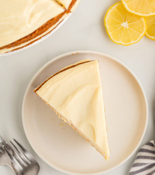 Overhead view of lemon cream cake slice on plate, with remaining cake and slices of lemon in background