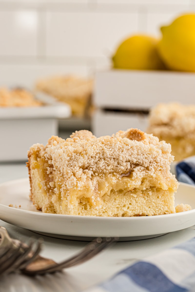 Lemon yogurt crumb cake piece on plate with rest of cake and lemons in background