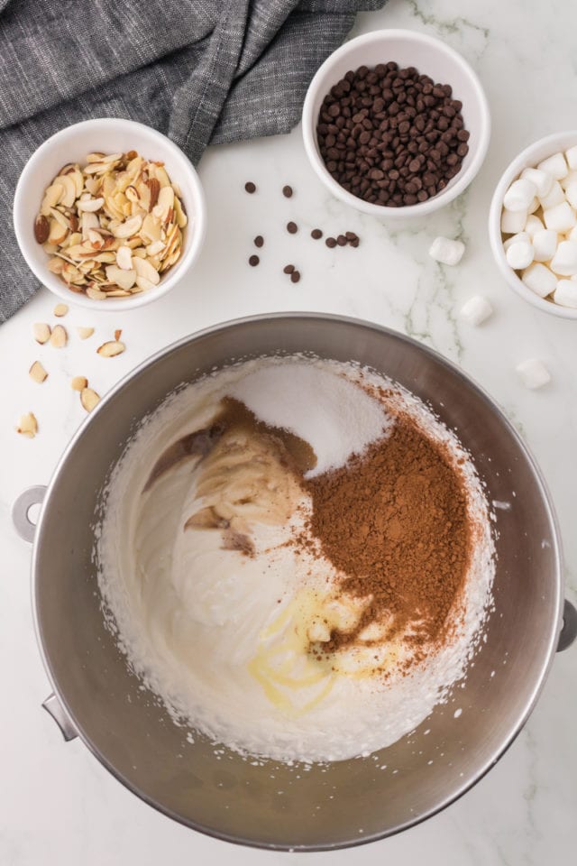 overhead view of cocoa powder, sugar, vanilla extract, and almond extract added to partially whipped cream in a metal mixing bowl