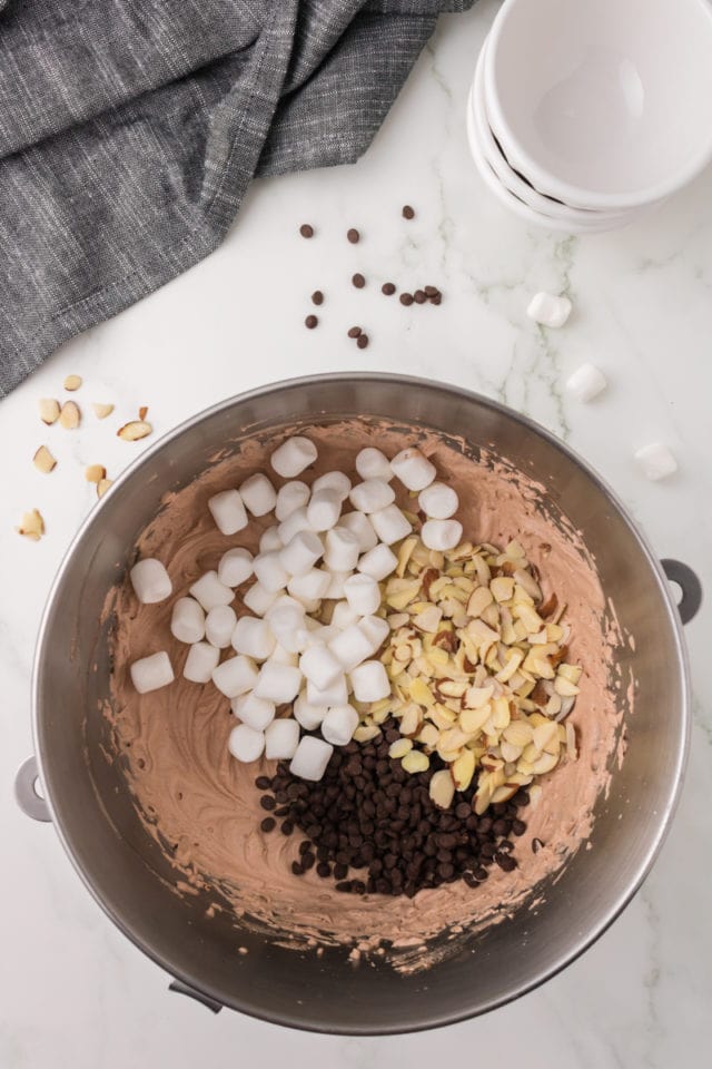 overhead view of marshmallows, chocolate chips, and almonds added to chocolate ice cream mixture in a metal mixing bowl