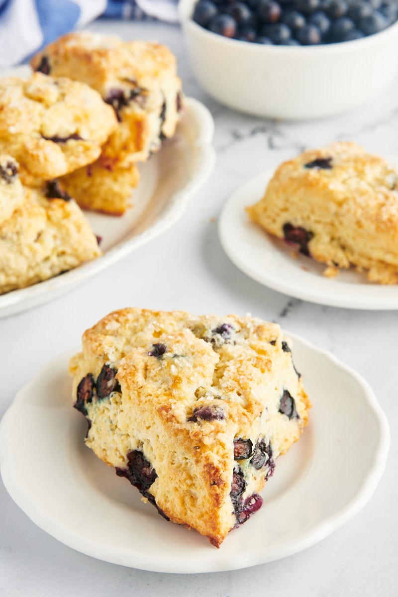 a blueberry scone on a white plate with more scones and a bowl of blueberries in the background