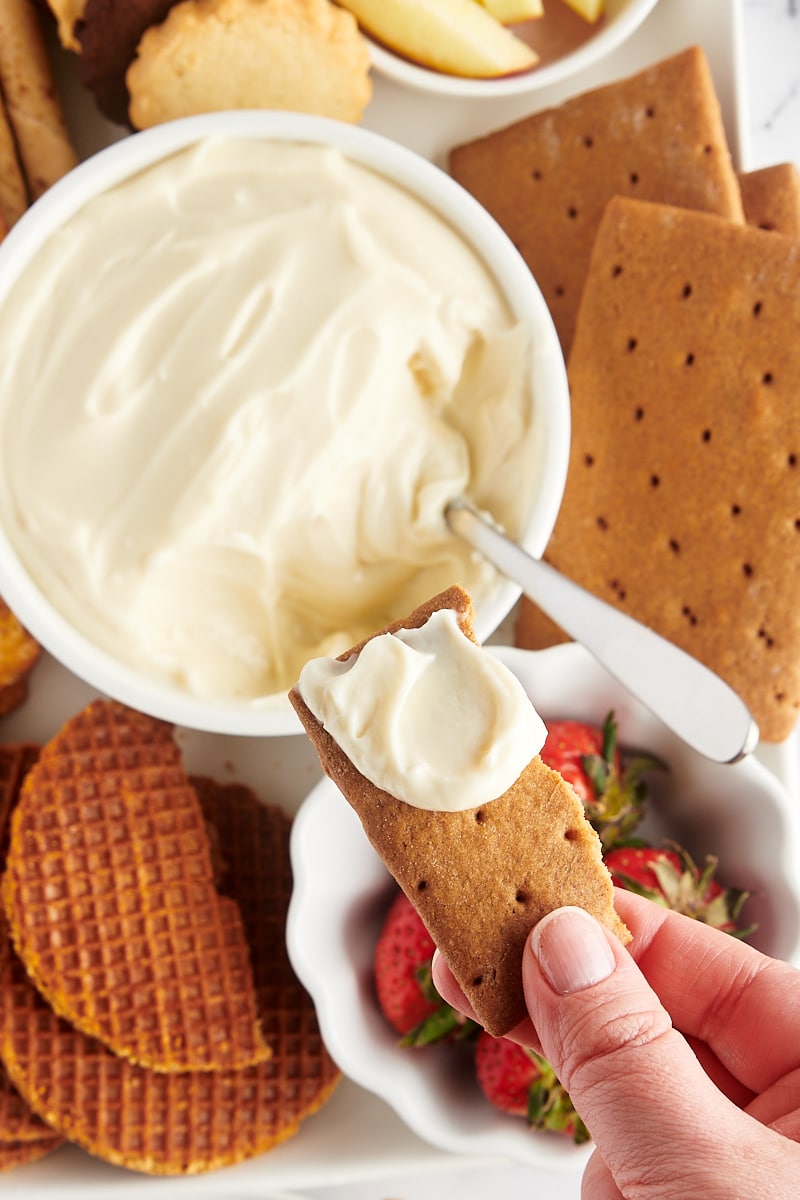 Overhead view of cheesecake dip in bowl with hand holding graham cracker piece