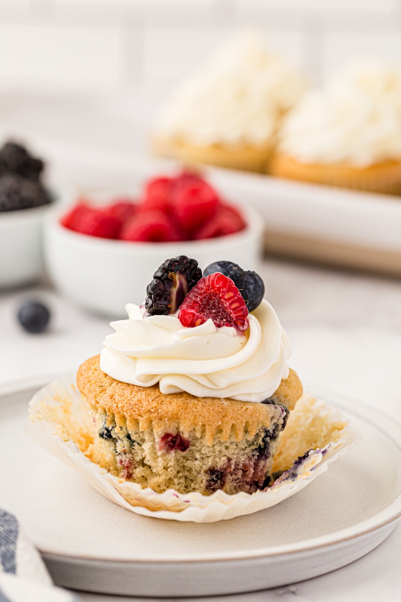 a berry cupcake on a white plate with more cupcakes and berries in the background