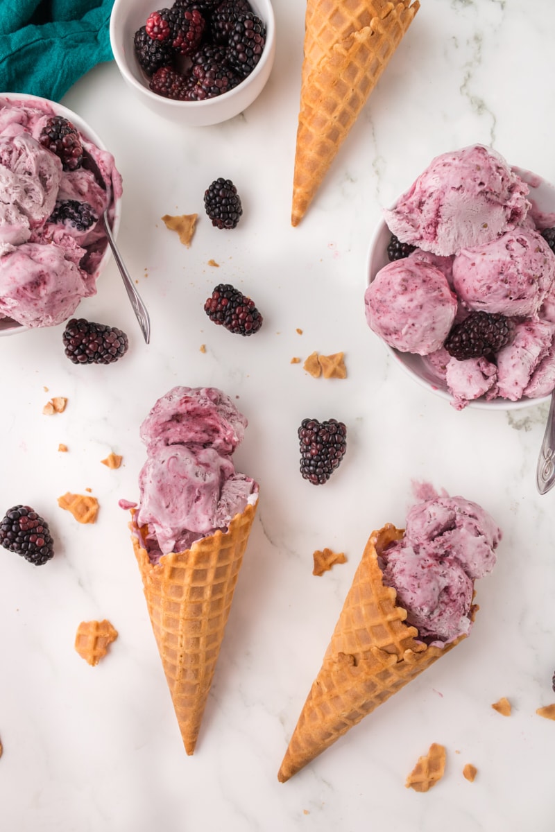 two cones and two bowls of blackberry ice cream on a marble surface