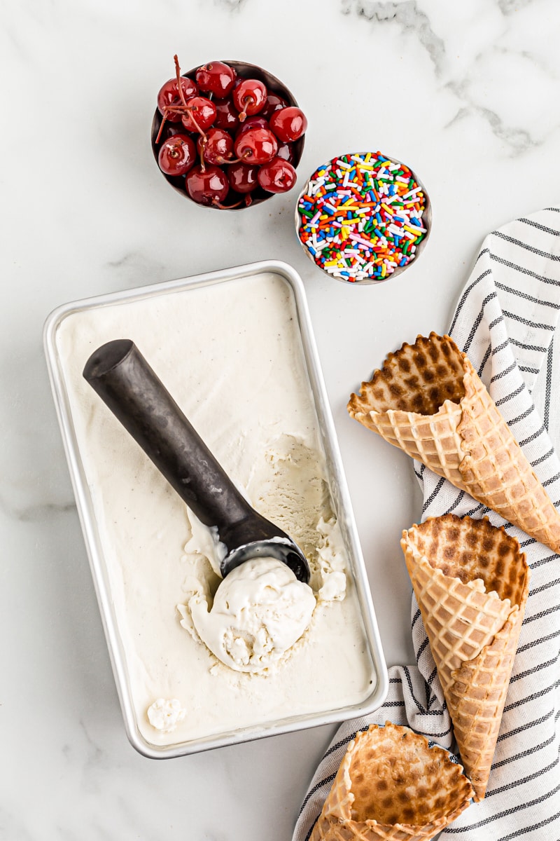 Overhead view of loaf pan of no-churn vanilla ice cream set next to 3 waffle cones and bowls of cherries and sprinkles