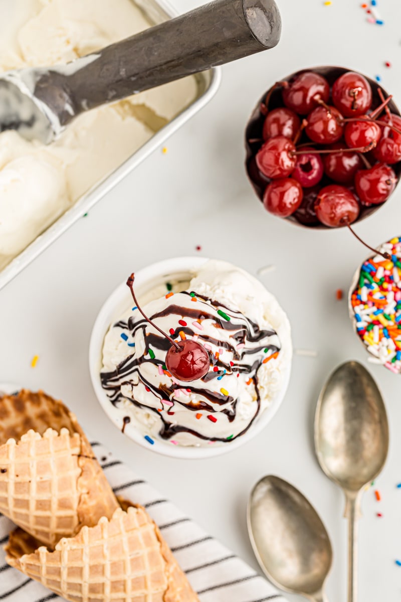 Overhead view of ice cream sundae in bowl, surrounded by vanilla ice cream, cherries and sprinkles in bowls, and ice cream cones