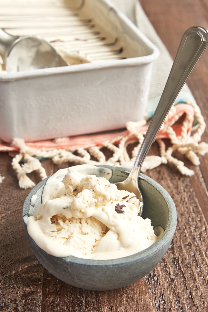 Partially melted stracciatella ice cream in gray bowl with spoon, with pan of ice cream in background