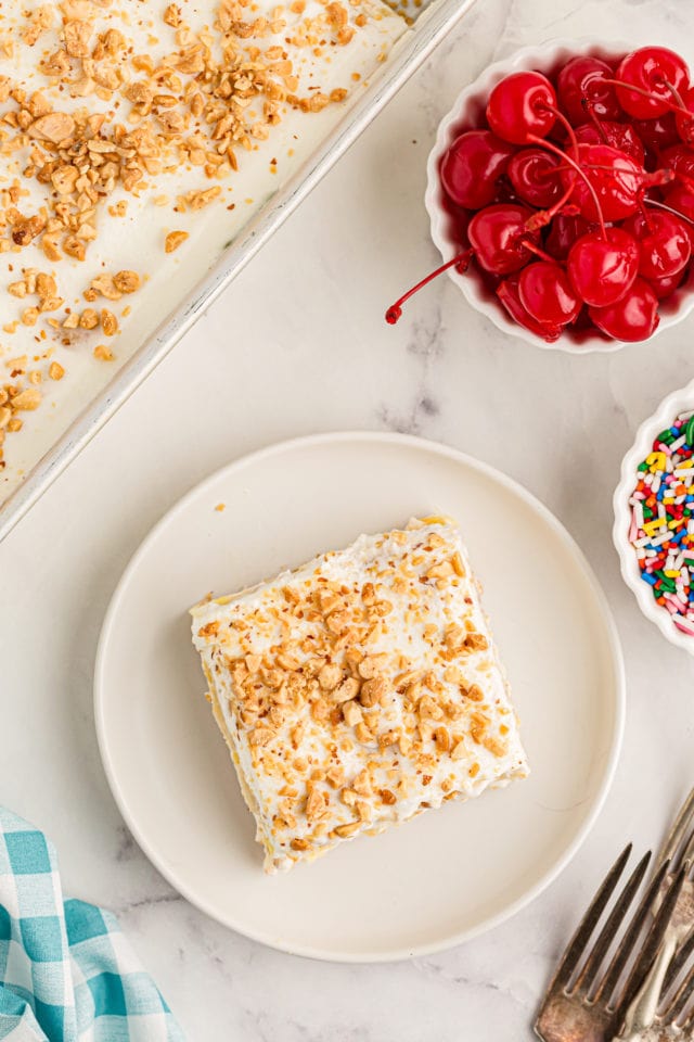 Overhead view of banana split cake on plate with bowls of cherries and sprinkles