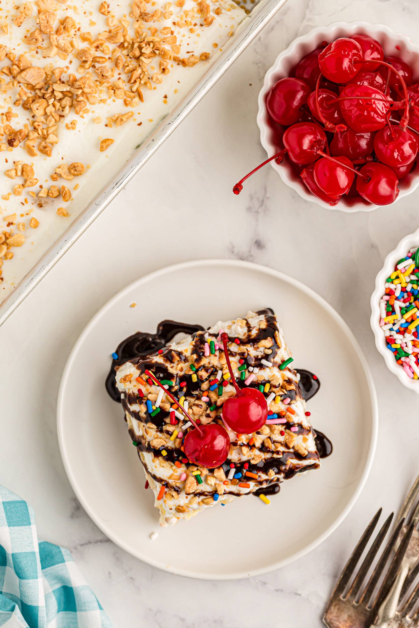 Overhead view of banana split cake on plate with bowls of cherries and sprinkles