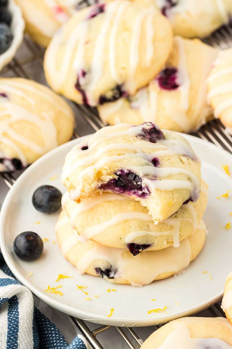 a stack of three lemon blueberry cookies on a white plate with more cookies surrounding