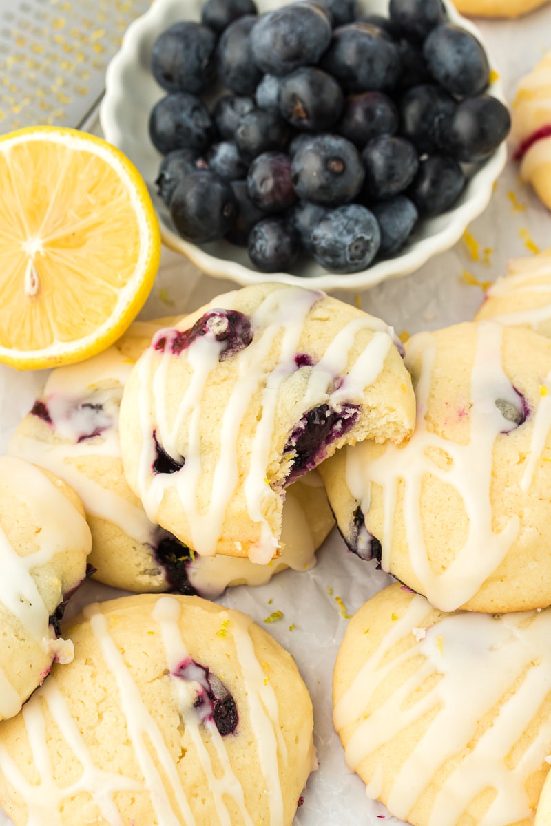 a lemon blueberry cookie with a bite missing on top of more cookies with a bowl of blueberries in the background