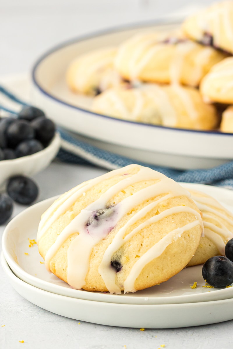 two lemon blueberry cookies on a white plate with a large white plate of cookies in the background