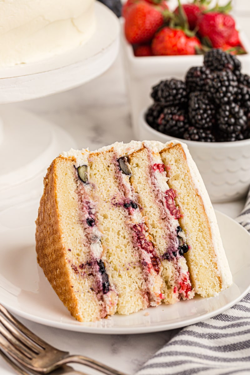 Slice of chantilly cake on its side on a white cake, with bowls of berries in background