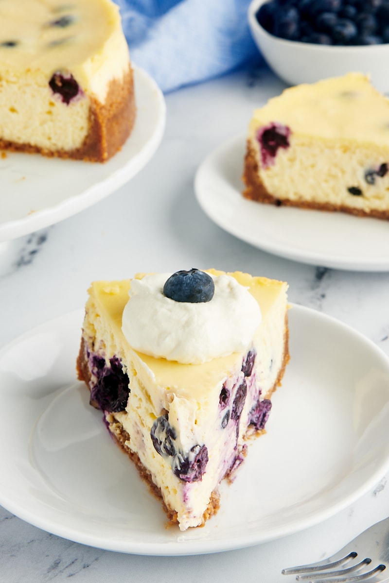 a slice of blueberry cheesecake on a white plate with two more servings in the background