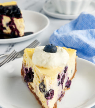 a slice of blueberry cheesecake on a white plate with another slice and a bowl of blueberry compote in the background