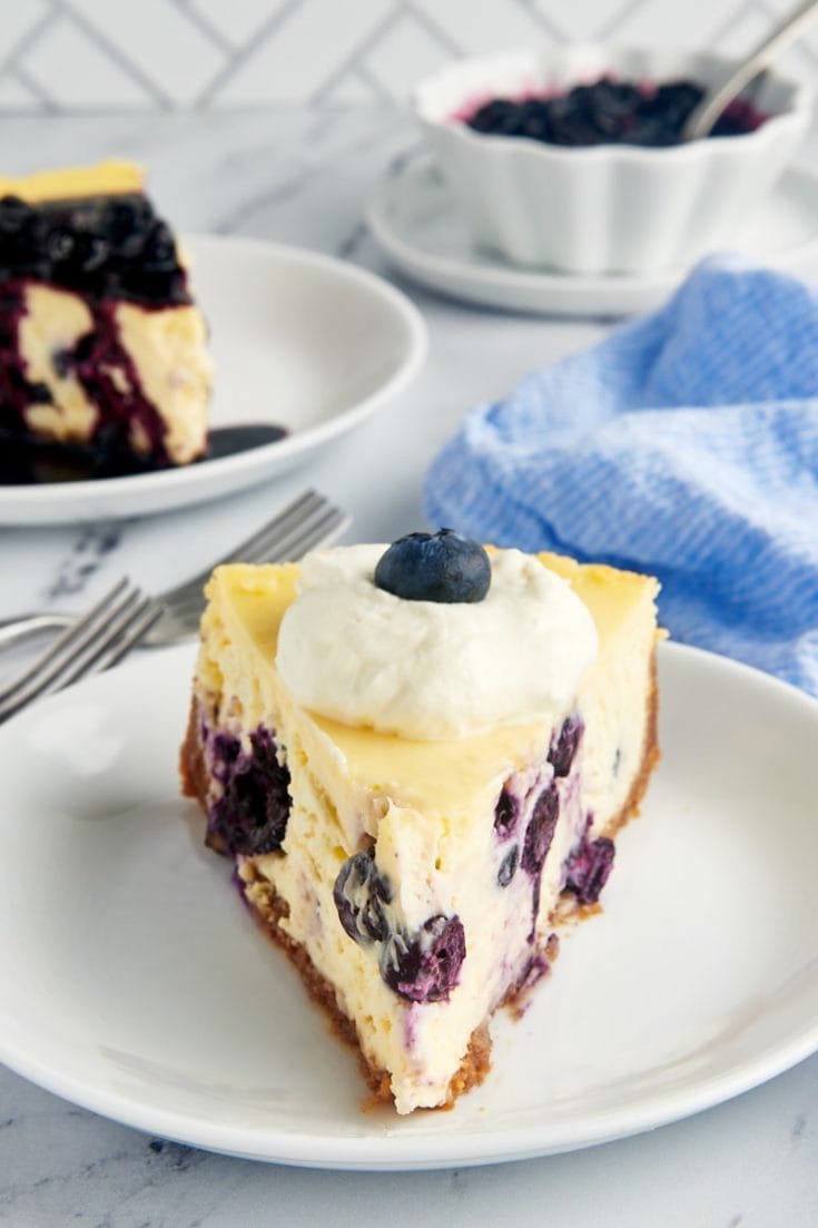 a slice of blueberry cheesecake on a white plate with another slice and a bowl of blueberry compote in the background