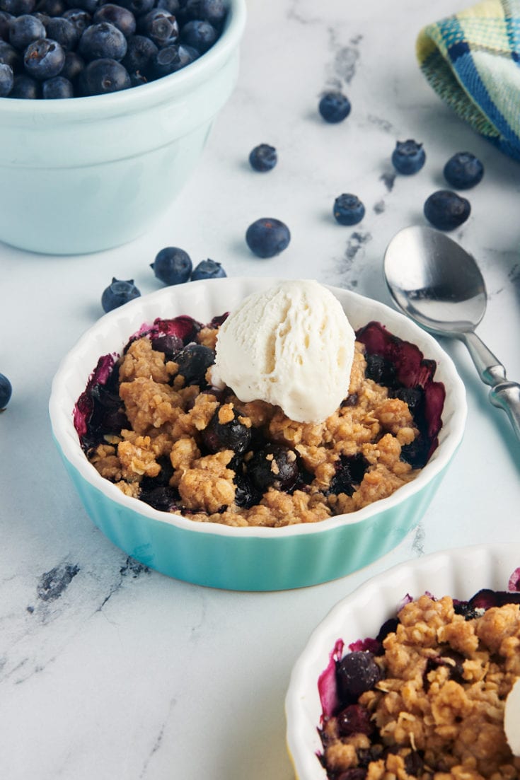 Two small baking dishes of blueberry crumble with bowl of blueberries in background