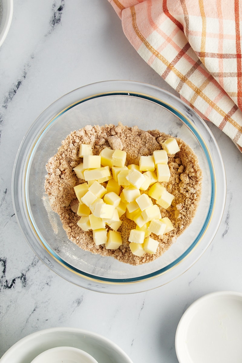 Cubed cold butter being added to the dry crumb topping mixture in a glass bowl.