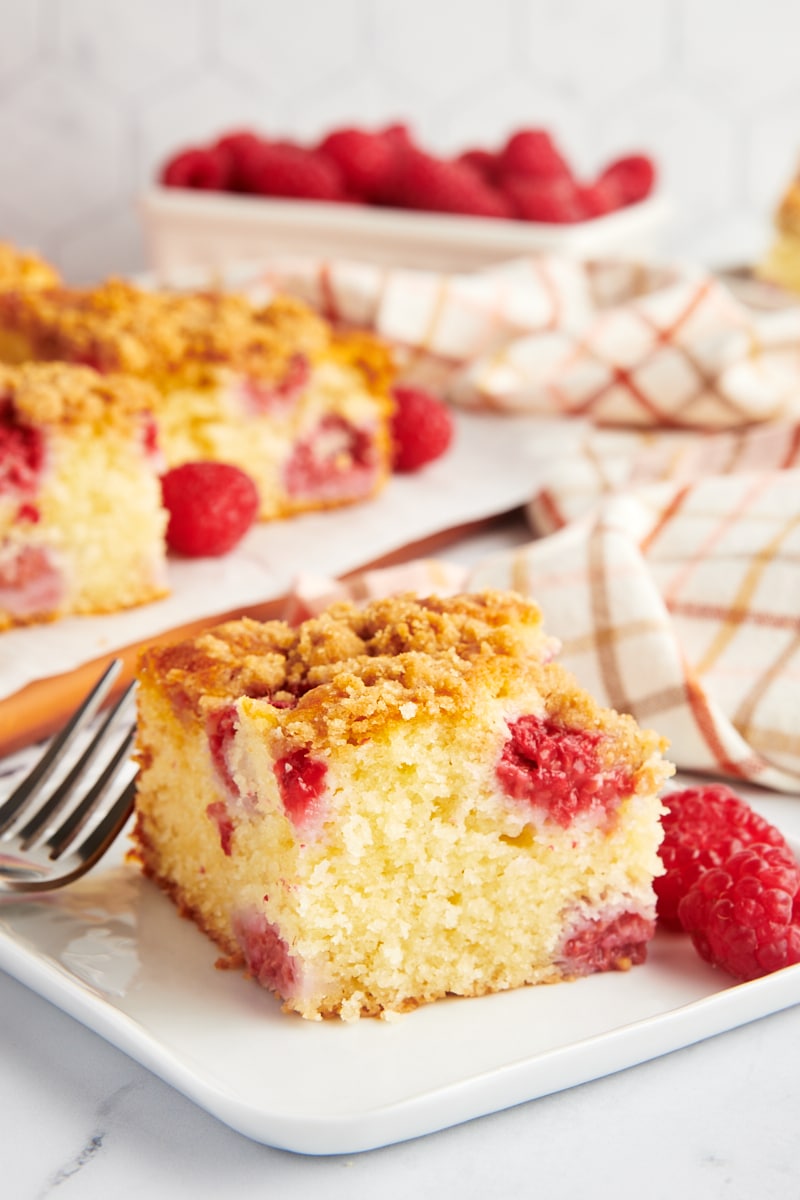 a slice of raspberry coffee cake on a square white plate with more cake and raspberries in the background